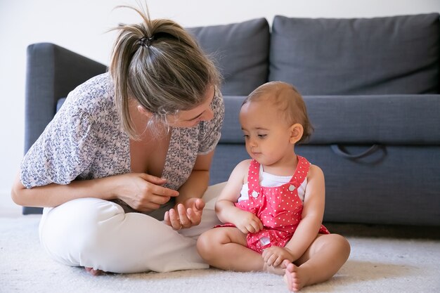 Happy little girl listening mom and sitting on carpet barefoot. Blonde mother sitting cross-legged and talking to daughter. Lovely serious infant looking at mum hands. Weekend and motherhood concept