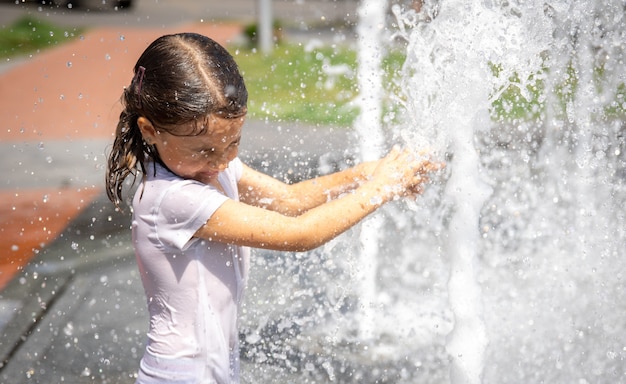 街の噴水のしぶきの中の幸せな少女は楽しんで、暑さから逃げます。