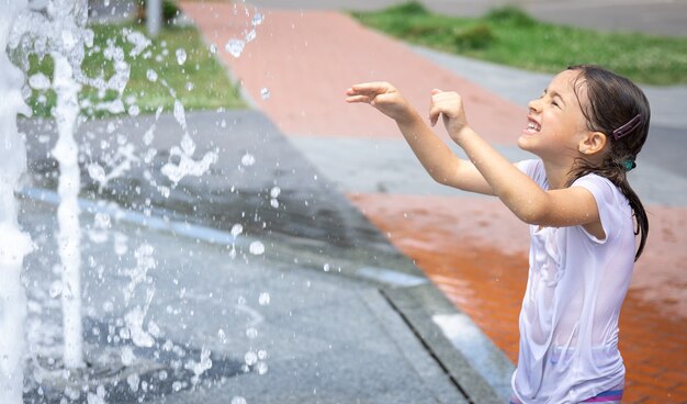 Happy little girl among the splashing water of the city fountain has fun and escapes from the heat.
