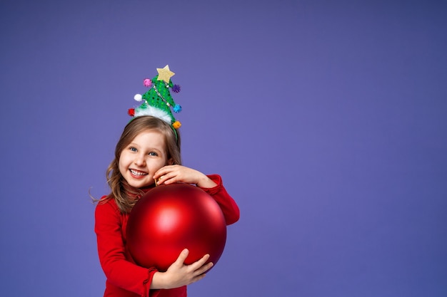 Happy little girl with decorated headband holds christmas red ball on purple in studio.