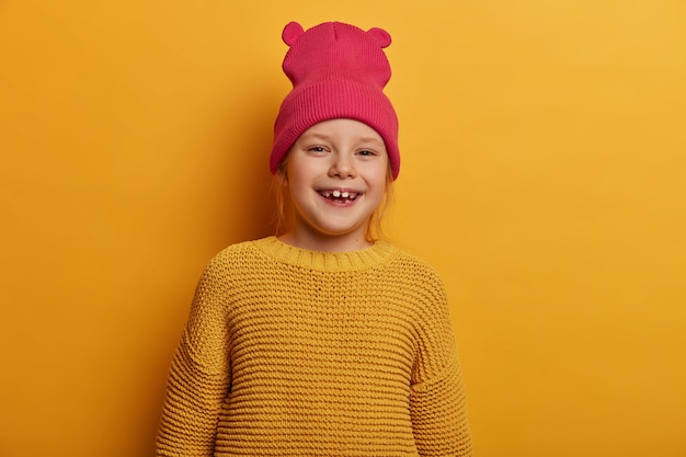 Счастливая маленькая девочка с довольным выражением лица смотрит прямо, позитивно улыбается, выражает искренние эмоции, оптимистично, носит розовую шапку с ушками и вязаный желтый свитер, позирует в помещении
