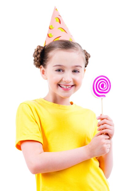 Happy little girl in yellow t-shirt and party hat holding colored candy - isolated on white.