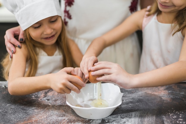 Счастливые маленькие девочки разбивают яйца в миску над кухонной столешницей