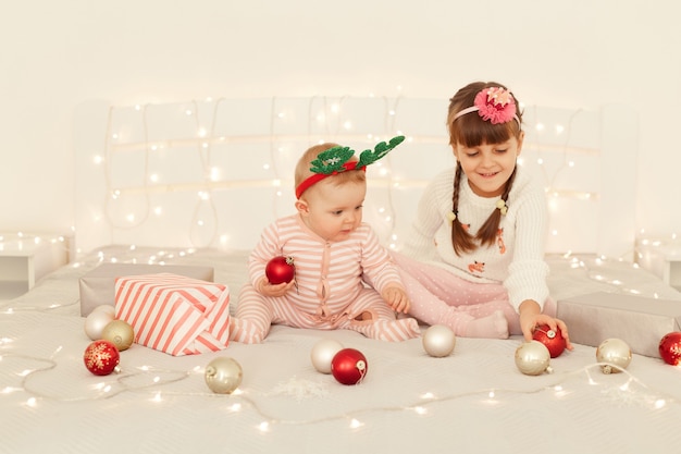 Happy little girls siblings wearing casual attires and party props, playing together while sitting on bed with Christmas decorations, celebrating New year eve.