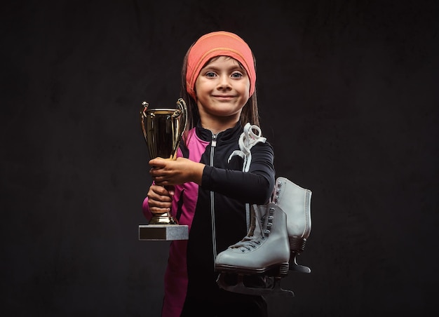 幸せな小さなスケーターの女の子のチャンピオンは、勝者のカップとアイススケートを保持しています。暗いテクスチャの背景に分離されています。