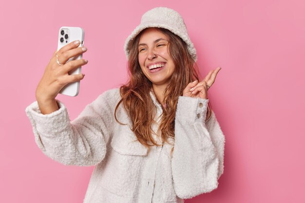 Happy long haired European woman makes peace gesture takes selfie on mobile phone smiles broadly dressed in fashionable winter clothes isolated over pink background enjoys video call with friend