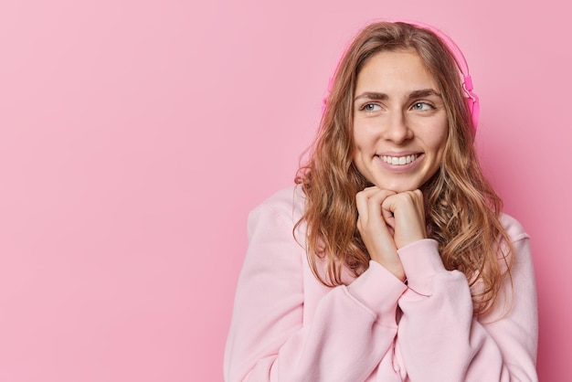 Happy long haired woman keeps hands near chin looks away with glad expression wears casual sweatshirt isolated over pink background blank copy space for your advertising content enjoys music