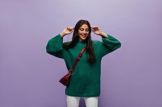 Happy longhaired woman in green sweater and white pants poses on isolated Brunette lady in woolen top smiles on purple background