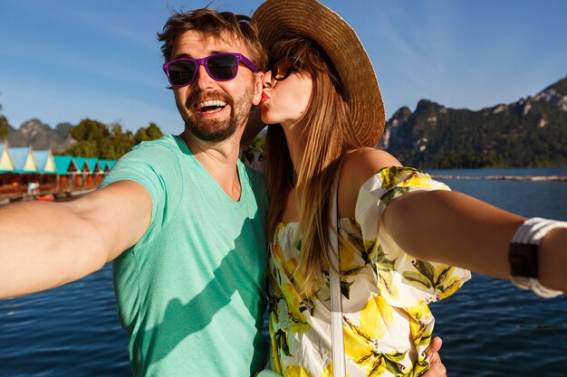 山や湖での休暇で自分撮りをする幸せな素敵なカップル、夏の明るい服の帽子とサングラス、キスと一緒に楽しい。