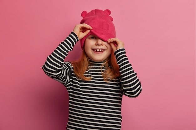 Free photo happy lovely girl preschooler looks from under hat, plays hide and seek, wears casual striped jumper, isolated over rosy pastel wall, has healthy skin, hears hilarious story, laughs positively