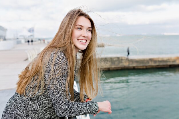 長い薄茶色の髪と大きな青い目が笑って海の近くを歩く幸せな素敵な女性