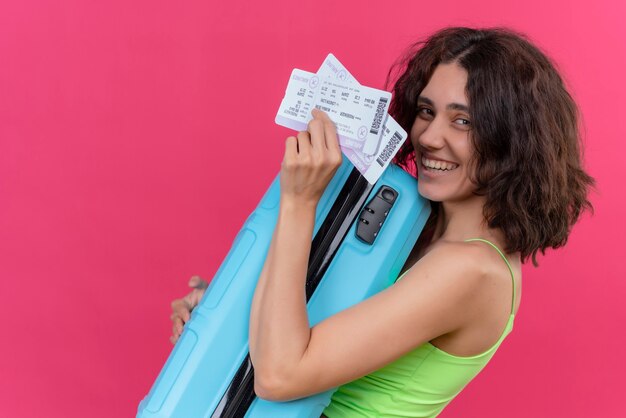 A happy lovely woman with short hair wearing green crop top showing plane ticket holding blue suitcase