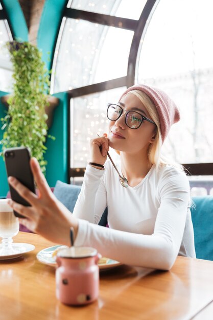 Happy lovely young woman in hat and glasses eating and making selfie with smartphone in cafe