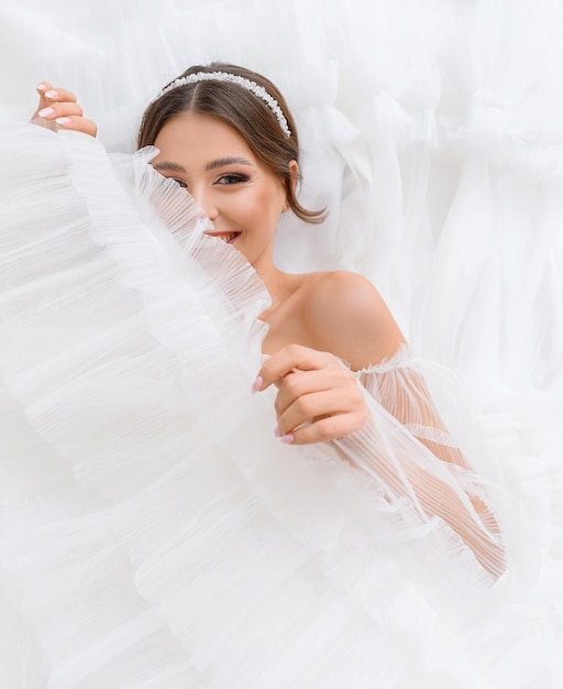 カメラを覗き込んでいる彼女の結婚式の白いふくらんでいるドレスの上に横たわっている幸せな素敵な若い女性完璧なヌードメイクとジュエリーで笑顔の花嫁の女の子ウェディングドレスのコンセプト愛らしい花嫁のウェディングサロン