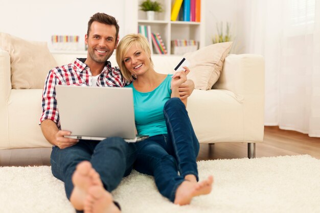 Happy loving couple sitting on the floor and using laptop and showing credit card
