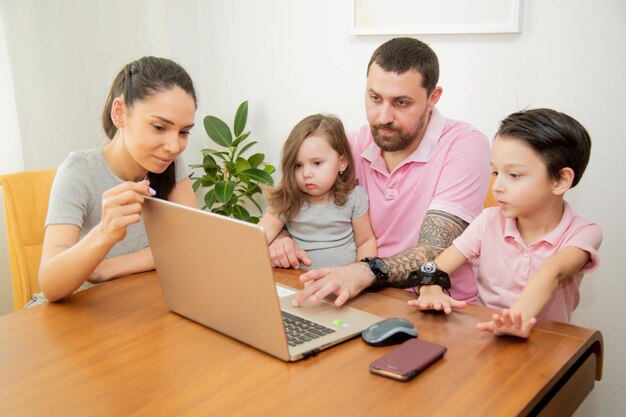 幸せな愛情のある家族のカップルの両親と小さなかわいい子供たちの息子と娘が一緒にテーブルに座っている父は家で働く忙しい顔でラップトップに取り組んでいますコンセプト