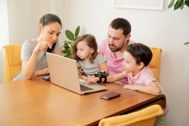 Счастливая любящая семейная пара, родители и маленькие милые дети, сын и дочь, сидят за столом вместе, отец работает на ноутбуке с занятым лицом, работая дома