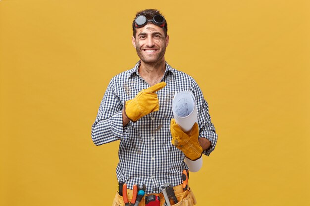 Счастливый рабочий по техническому обслуживанию или ремонтник с грязным лицом в защитных очках, перчатках и поясе с инструментами, держащими план, стоящим у желтой пустой стены, указывая на copyspace