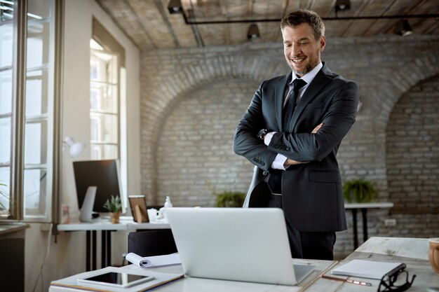 Happy male entrepreneur standing with arms crossed while reading something on a computer in the office