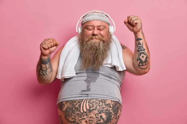 Free photo happy male fatso enjoys workout with music, raises hands and dances, has sweaty body, wears shirt and towel around neck, isolated on pink wall. chubby athlete happy to achieve great results