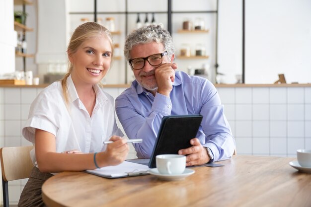 Happy male and female colleagues of different ages sitting at table at co-working, using tablet together, writing notes, looking at camera, smiling