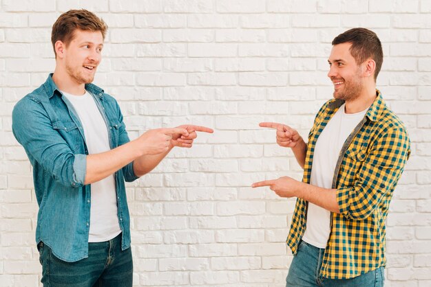 Happy male friends standing against white wall pointing their fingers to each other