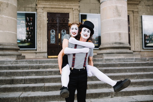 Happy male mime giving piggyback ride to female mime in front of staircase