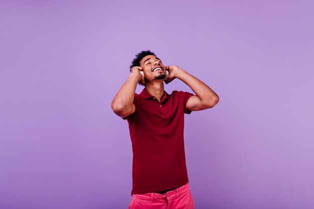 Happy male model listening music with eyes closed. Indoor shot of laughing carefree man in red t-shirt posing in headphones.