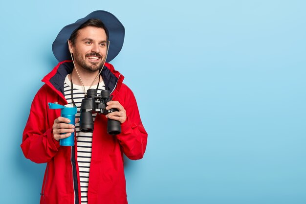 Happy male traveler explores new place, looks for something in distance with binoculars, holds blue thermos with drink, wears casual outfit