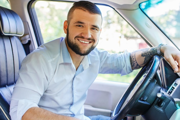 Happy male with black hair and beard, tattoes on his hand, dressed in white shirt and blue jeans shorts driving a car.