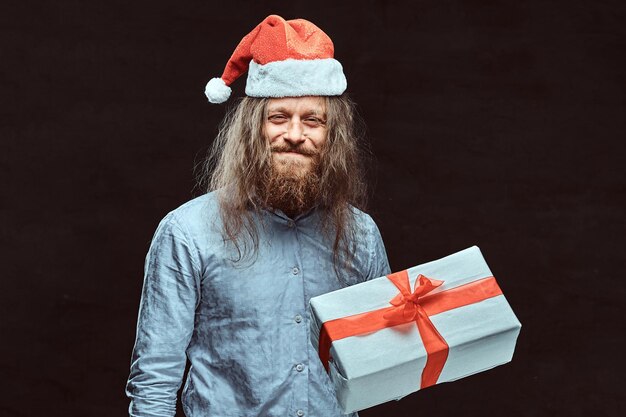 Счастливый мужчина с длинными волосами и бородой в синей рубашке и красной шляпе Санты держит подарочную коробку. Изолированные на темном фоне.