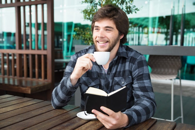 Счастливый человек пьет кофе и читает дневник в кафе на открытом воздухе