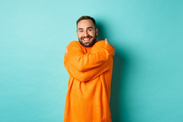 幸せな男は快適に感じ、暖かいセーターを着て抱きしめ、満足のいく笑顔で、明るいターコイズブルーの壁の上に立っています。