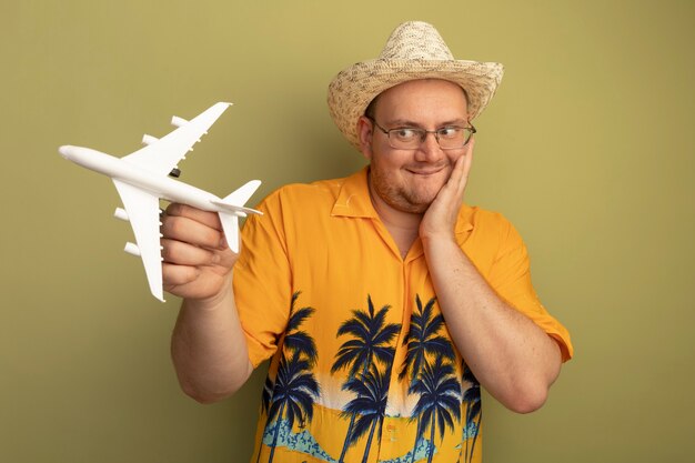 Happy man in glasses wearing orange shirt in summer hat holding toy airplane looking at it amazed standing over green wall