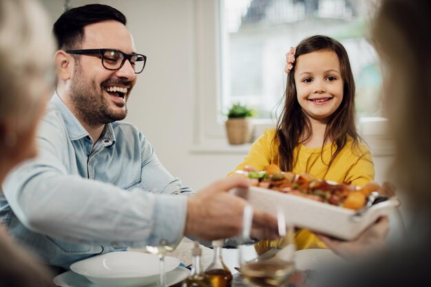 Happy man and his daughter serving food during lunch in dining room Focus is on little girl