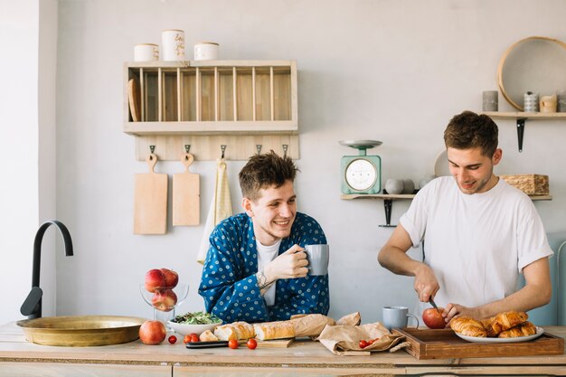 Happy man holding cup of coffee and his friend cutting apple on chopping board in kitchen