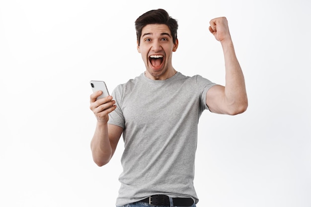 Free photo happy man holds smartphone and celebrating, winning online prize, shouts yes and rejoicing, triumphing from success, looks satisfied, white background