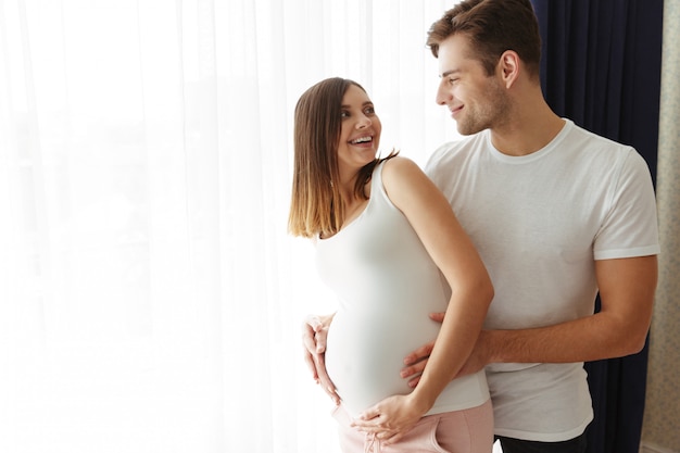 幸せな男は自宅で屋内で彼の素敵な妊娠中の妻を抱擁します。