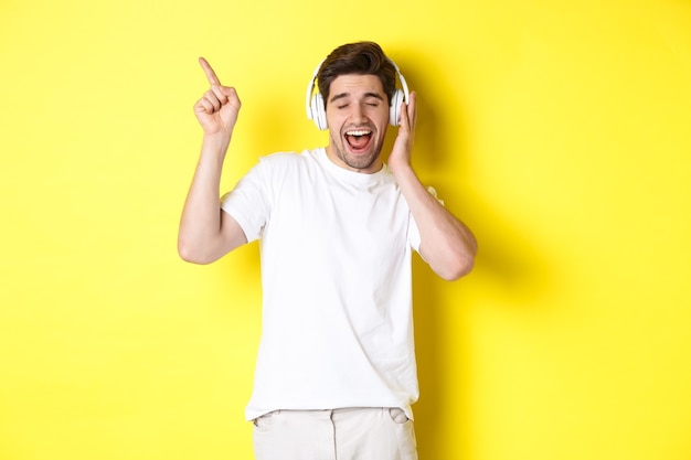 헤드폰을 끼고 음악을 듣고 있는 행복한 남자, 검은 금요일에 대한 프로모션 제안을 가리키는 손가락, 노란색 배경 위에 서 있는