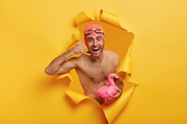 Happy man makes call gesture, wears swimhat and goggles, stands with naked torso, carries pink flamingo, being in good mood