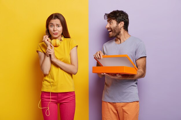 Happy man opens box with surprise, shows something to girlfriend who has unhappy puzzled look, frowns face, wears yellow t shirt and pink trousers, headphones around neck.