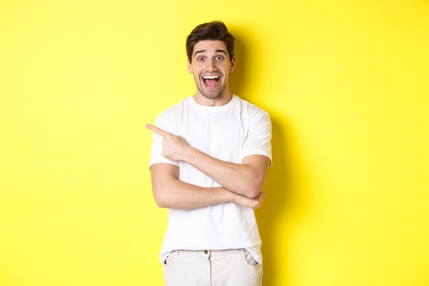 Happy man pointing finger left, showing advertisement on copy space, smiling amused, standing in white clothes against yellow background.