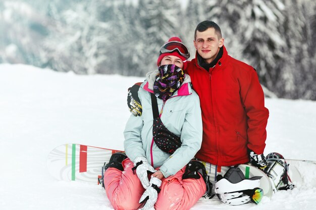 Happy man in red ski jacket and woman in blue one sit on their snowboards on mountain hill 