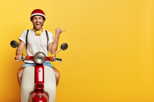 Happy man rides a scooter, shows direction away, points thumb right on blank space over yellow background, dressed in casual wear and helmet, uses headphones, has cheerful face expression