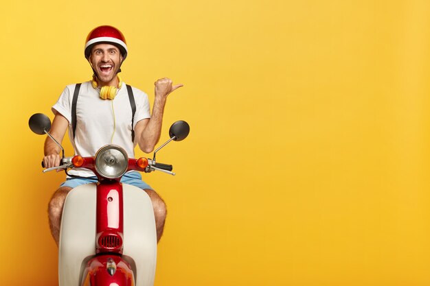 Счастливый человек едет на скутере, показывает направление, указывает большим пальцем прямо на пустое пространство на желтом фоне, одет в повседневную одежду и шлем, использует наушники, с веселым выражением лица