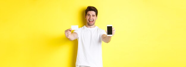신용 카드를 들고 서서 윙크하는 휴대폰 화면에서 좋은 온라인 제안을 보여주는 행복한 남자