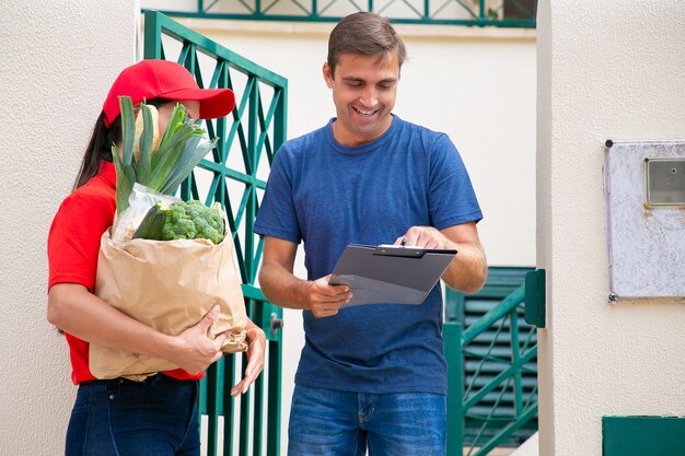 Happy man signing for receiving order from grocery store, holding clipboard and smiling. Postwoman in red uniform holding paper bag with vegetables. Food delivery service and post concept