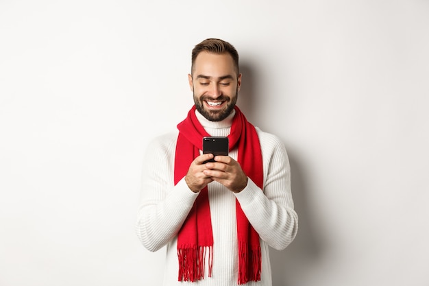 Happy man smiling while reading message on mobile phone, standing in winter sweater and red scarf against white background