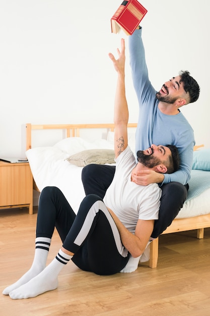 Happy man snatches the book from his boyfriend's hand sitting in the bedroom