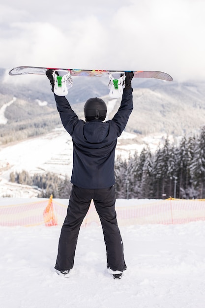Счастливый человек, стоящий со сноубордом над головой на вершине горного холма